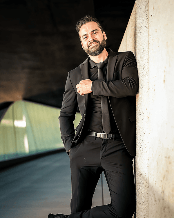 Eli Castson in a black suit leaning against concrete architecture