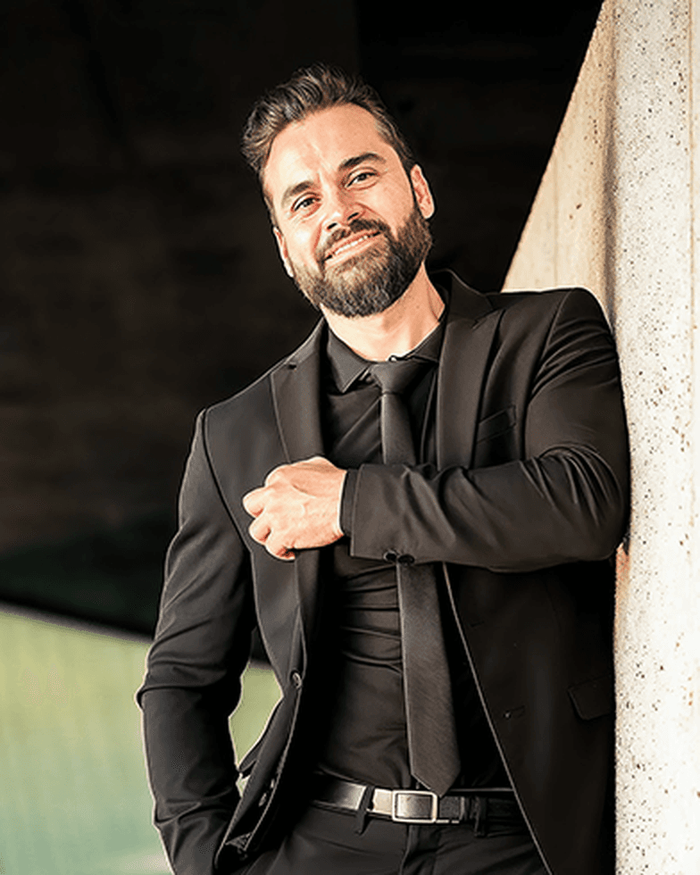 Eli Castson in a black suit leaning against concrete architecture